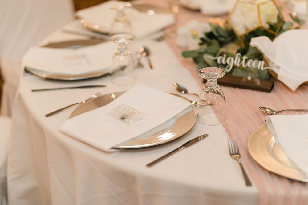 An elegant white and blush pink wedding reception table setting with pristine plates, silverware and decorative greenery.