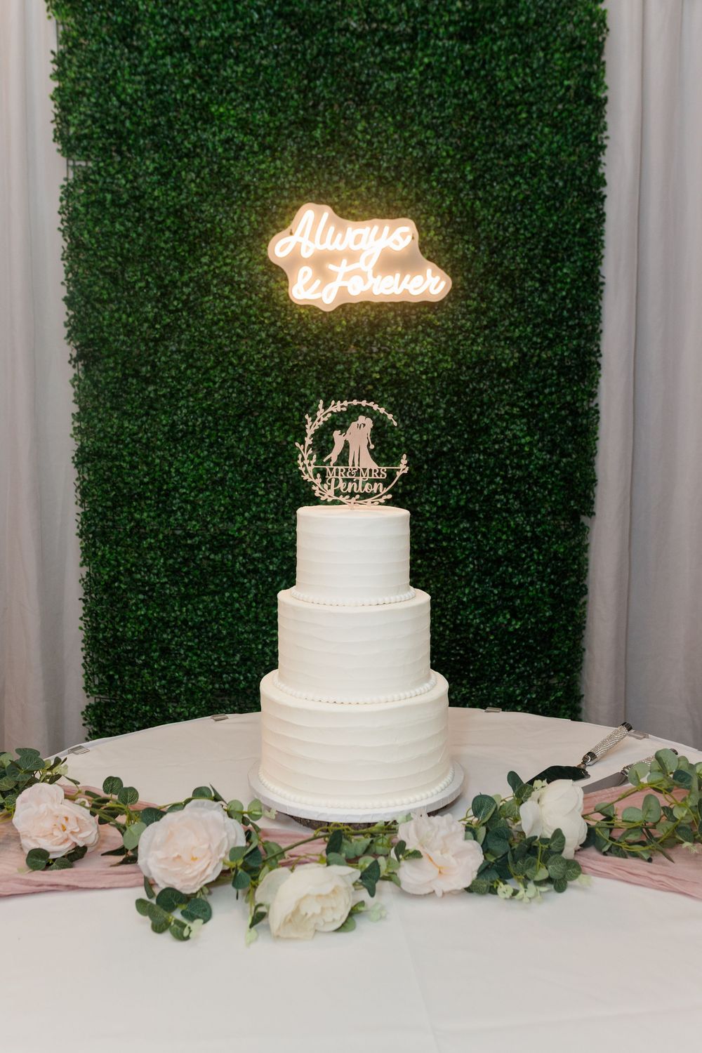 White wedding cake topped with glowing 'Always' neon sign on display with roses and greenery decorations.