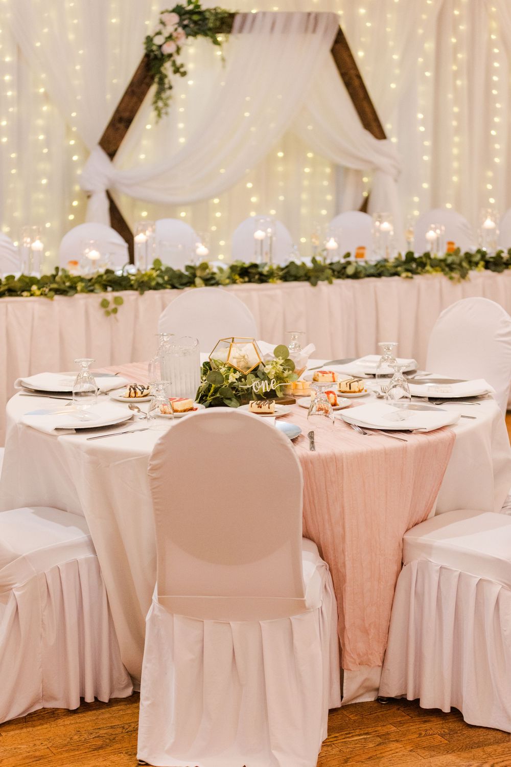 Elegant wedding reception table with white chair covers, fairy light backdrop and floral decorations in a romantic setting.