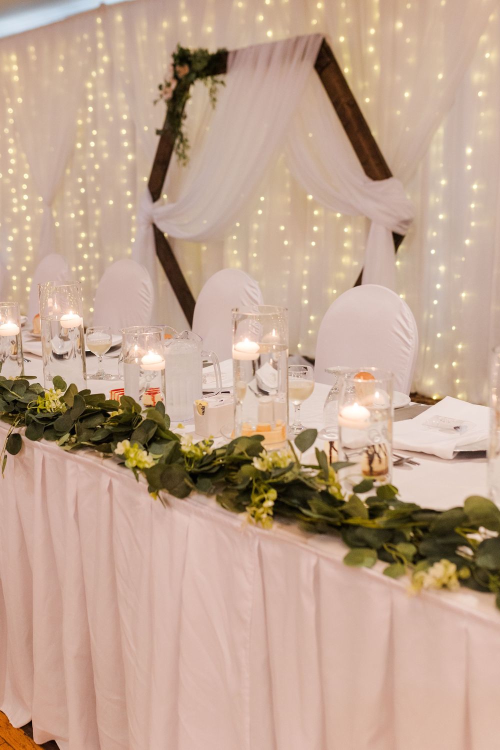 Wedding reception head table with white linens, floral garlands and twinkling fairy lights backdrop.