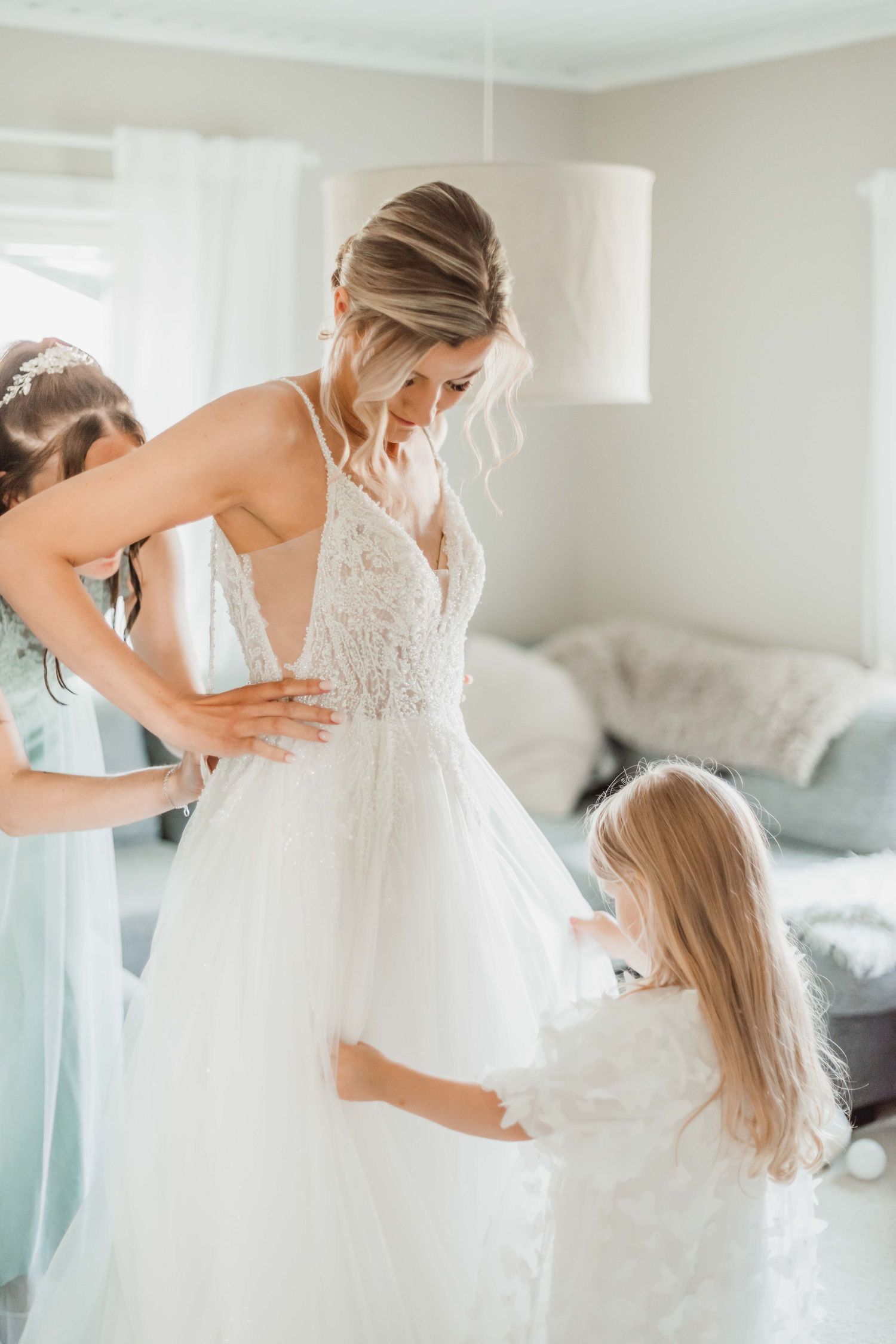 A heartwarming scene shows a young attendant in light blue helping adjust a bride's elegant white wedding gown.