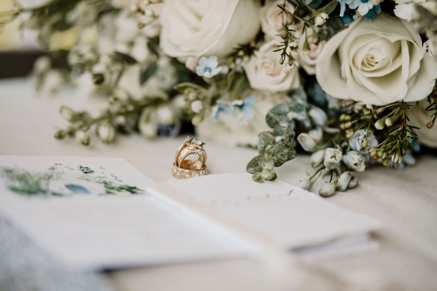 Wedding rings displayed next to white roses and delicate floral arrangements on a white surface.