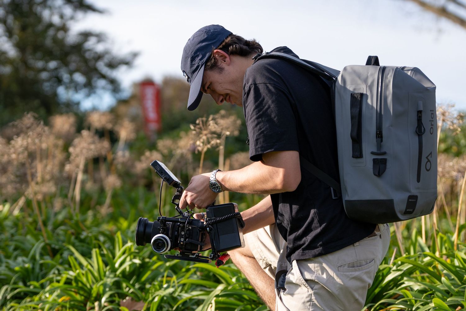 A person in a baseball cap films with a professional camera while wearing a backpack in an outdoor garden setting.