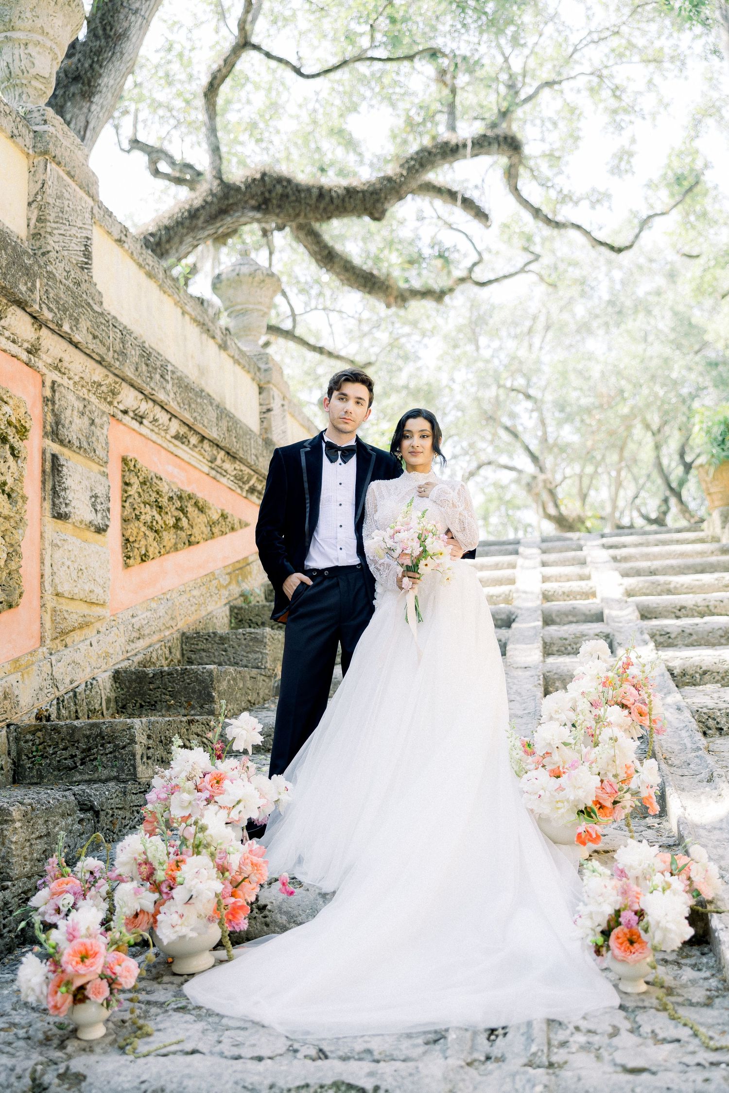 A romantic wedding couple stands on stone steps adorned with pink and peach floral arrangements under mossy oak trees.
