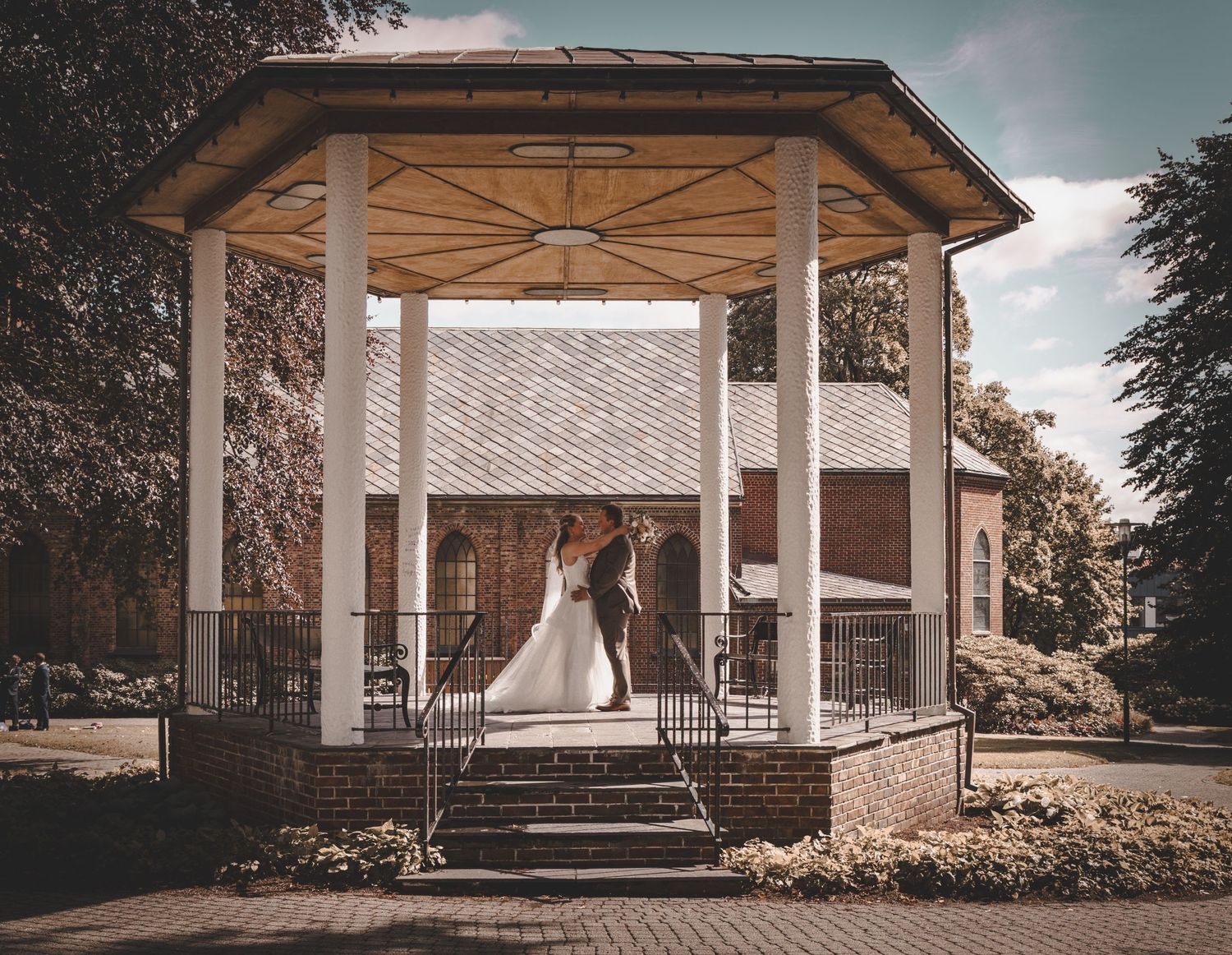 Bride and groom share tender kiss under elegant white gazebo in sunlit park setting.