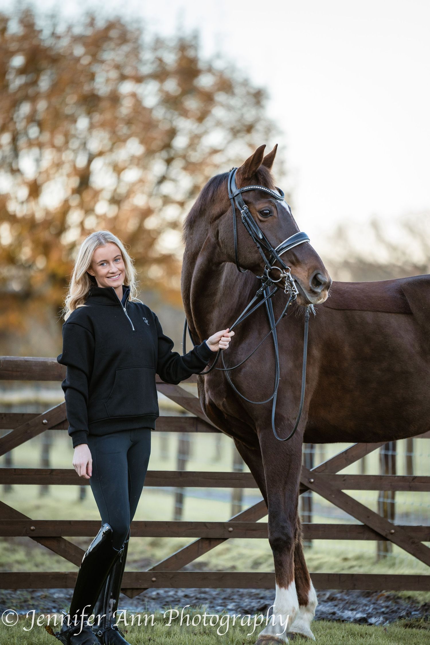A woman in black stands with a dark brown horse at a wooden fence during golden hour at a farm.