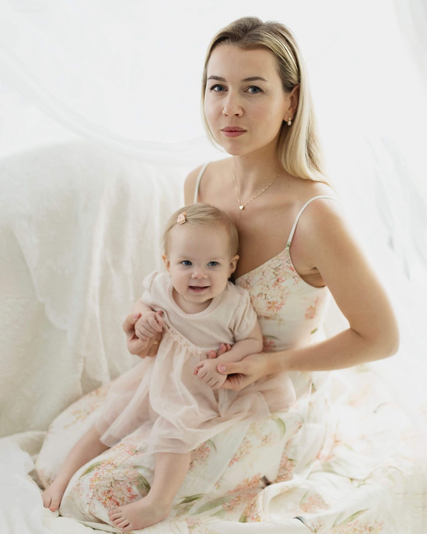Mother and baby wearing matching pink dresses pose together in a bright white bedroom setting with soft natural light.