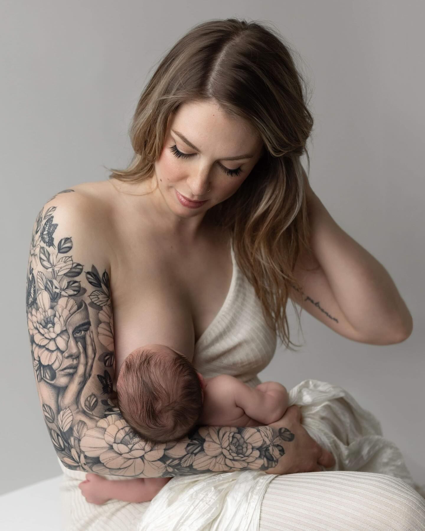 Person with floral arm tattoo nurses baby while reclining on white bedding in intimate monochromatic portrait.