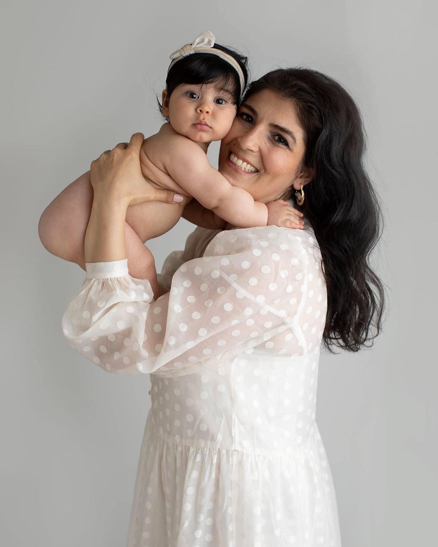 A mother and child share tender moments in matching white dresses against a light studio background.