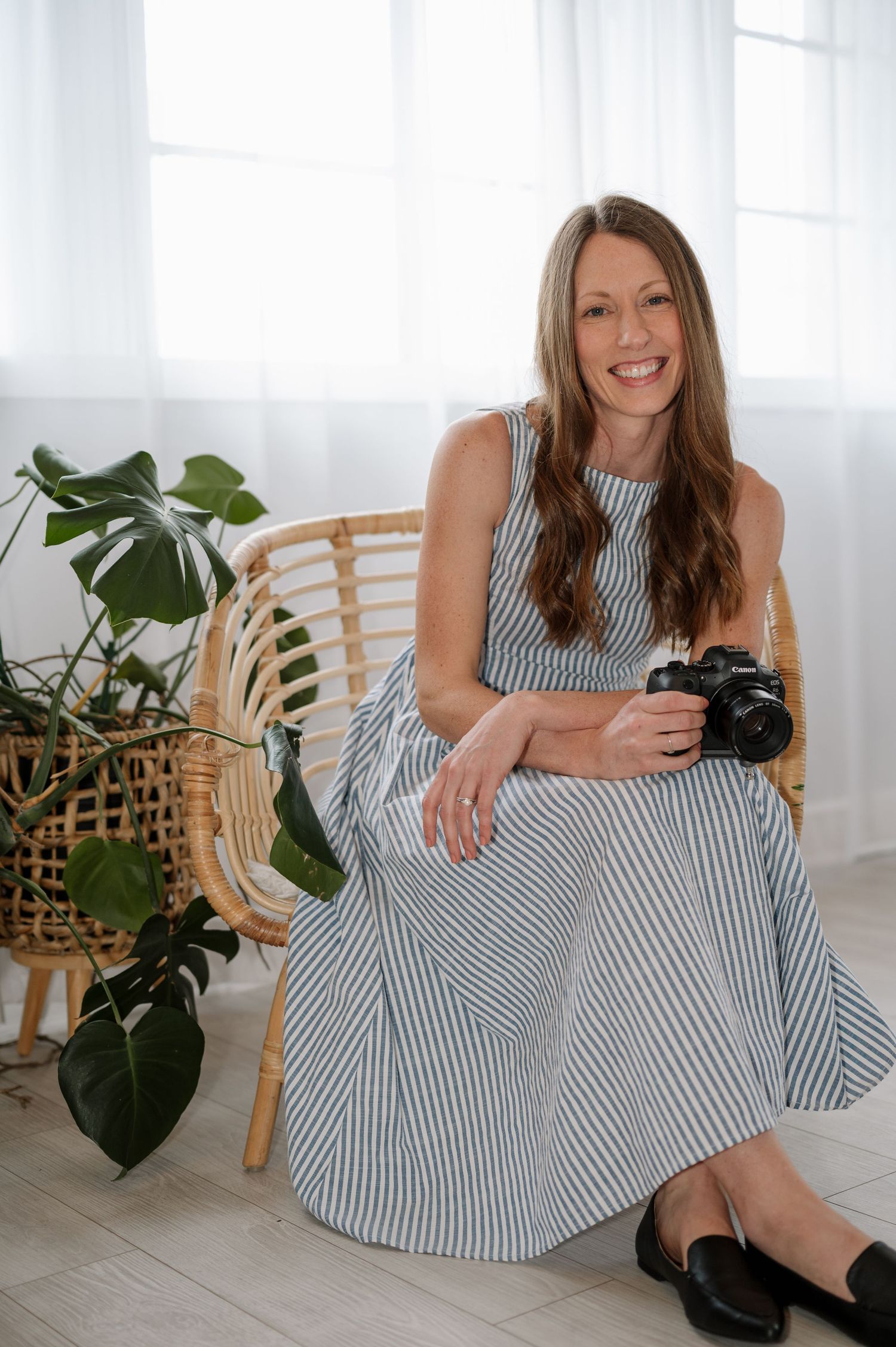 Professional photographer in striped dress sits with camera in rattan chair surrounded by plants.