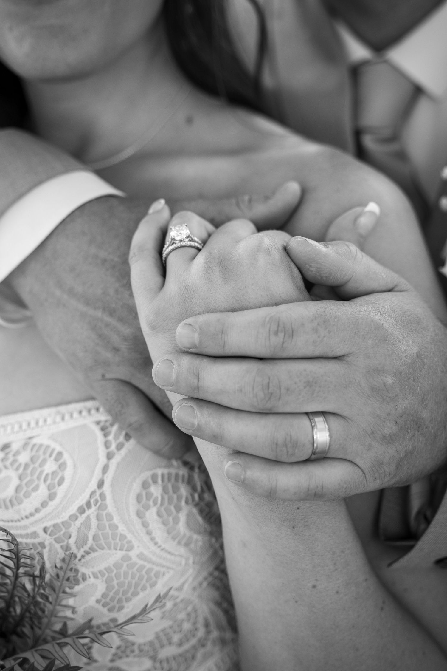 Black and white close-up of hands clasped together showing wedding rings and lace dress details.