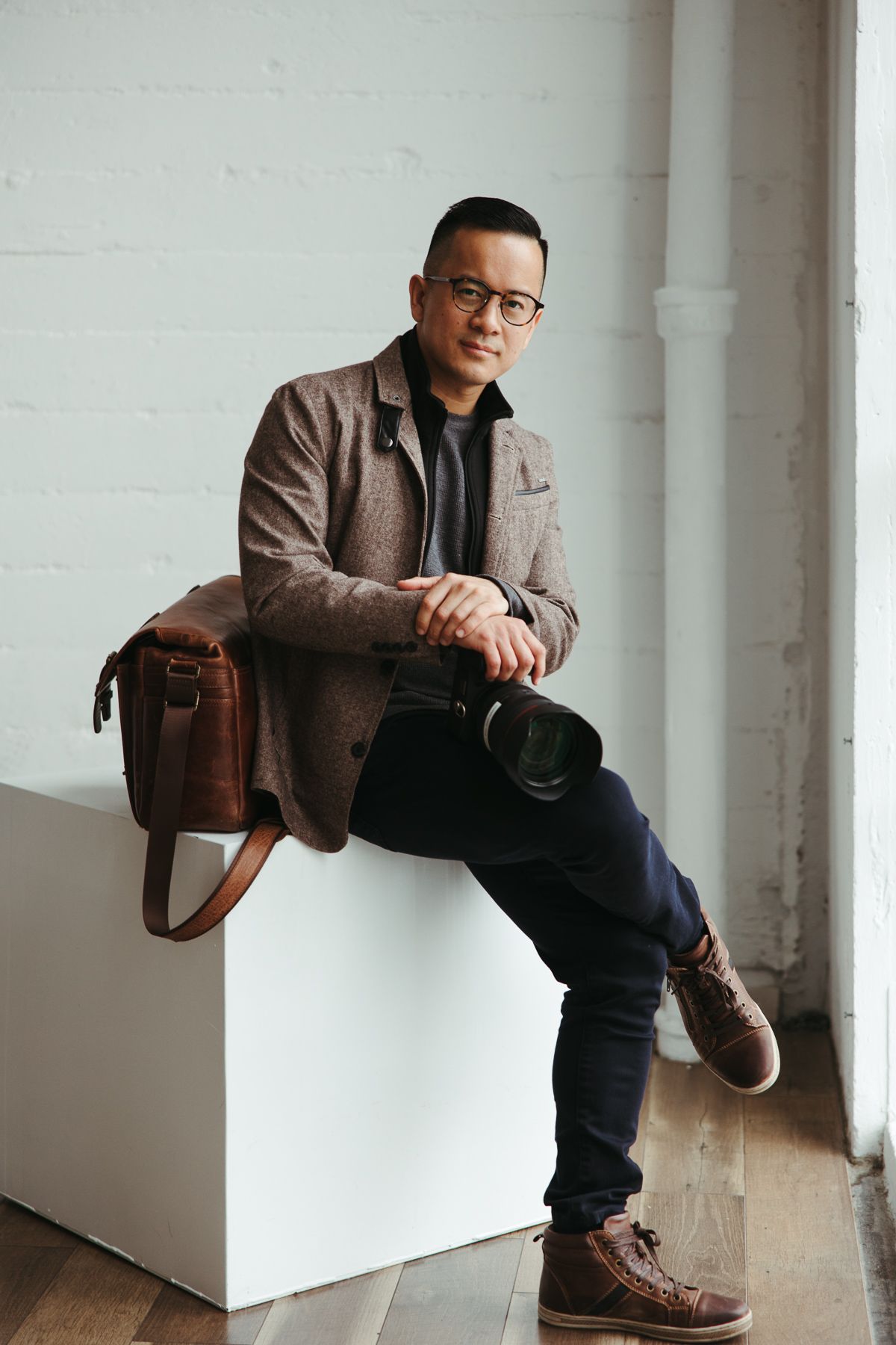 Stylish man in glasses and tweed blazer sitting on white cube with leather bag and hat in modern studio.