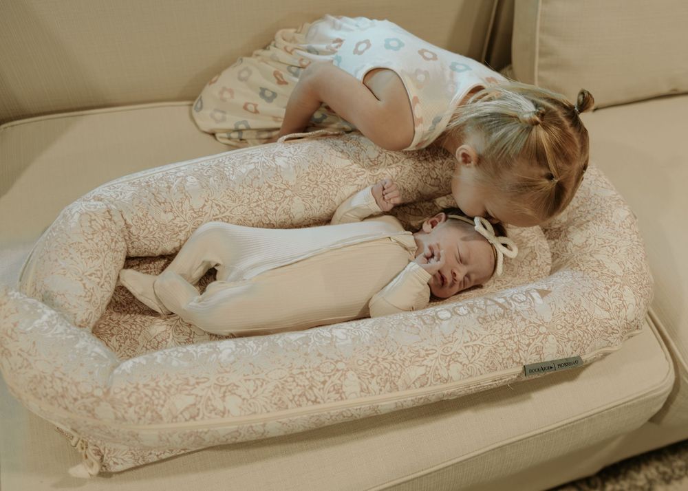 A baby lounges on a plush beige lounger pillow wearing white clothing.