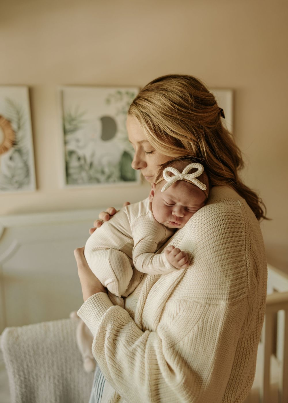A tender moment shows someone cradling a sleeping newborn wearing a bow headband in a nursery.