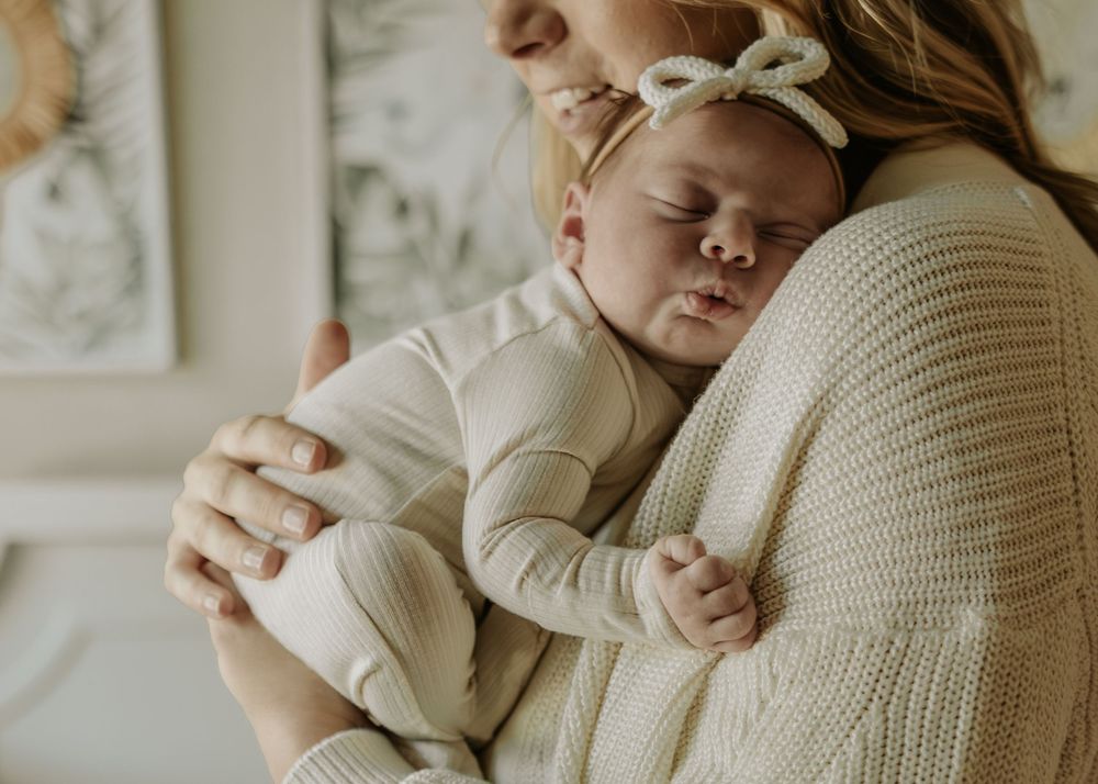 Sleeping infant in cream outfit with bow headband being held against knit sweater.