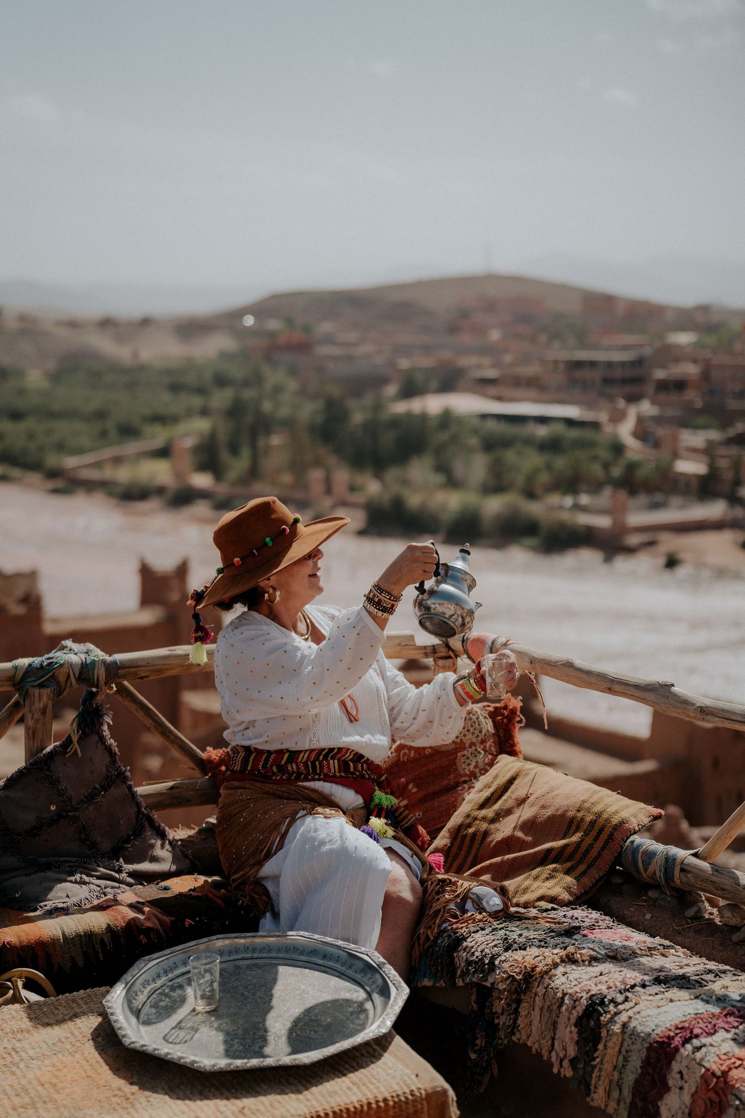 Person in white blouse and hat pours tea on cushioned outdoor terrace overlooking desert landscape.