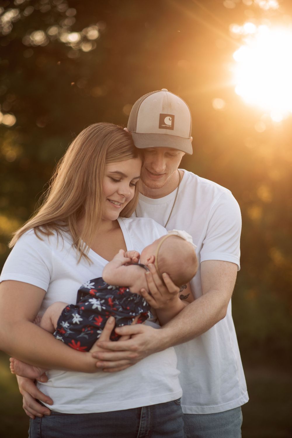 Young parents in white shirts tenderly holding newborn baby in golden backlit outdoor setting.