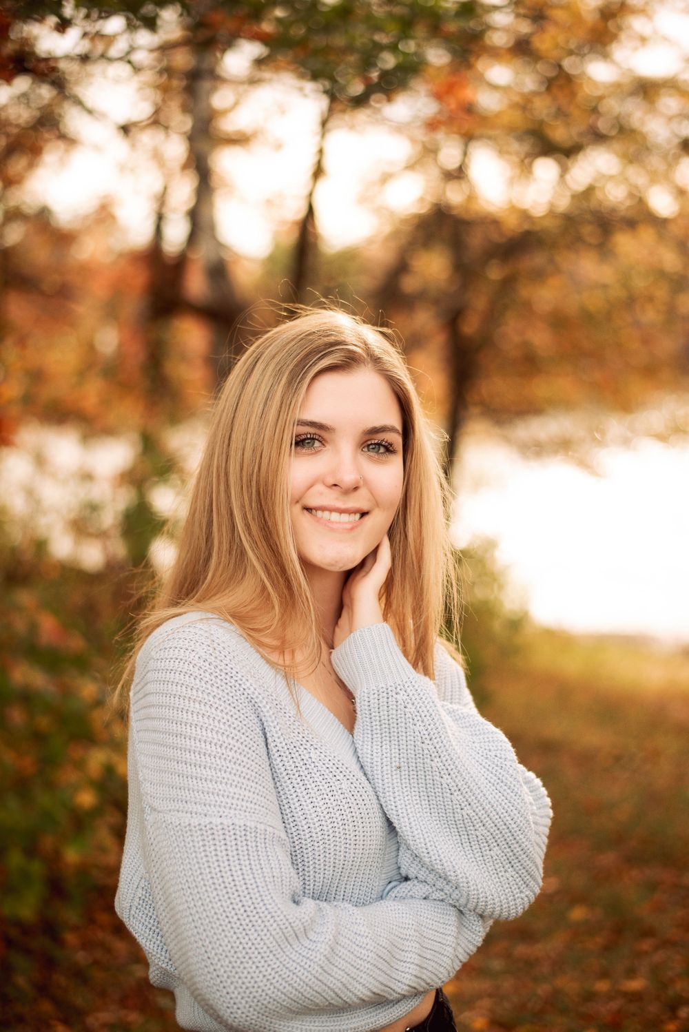 Smiling blonde teen girl in light blue sweater posing in autumn forest for senior portrait.