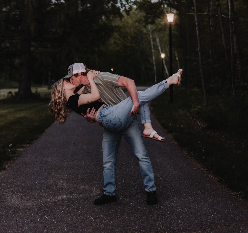 Couple sharing a romantic dip kiss on a tree-lined driveway at dusk with soft lamplight.