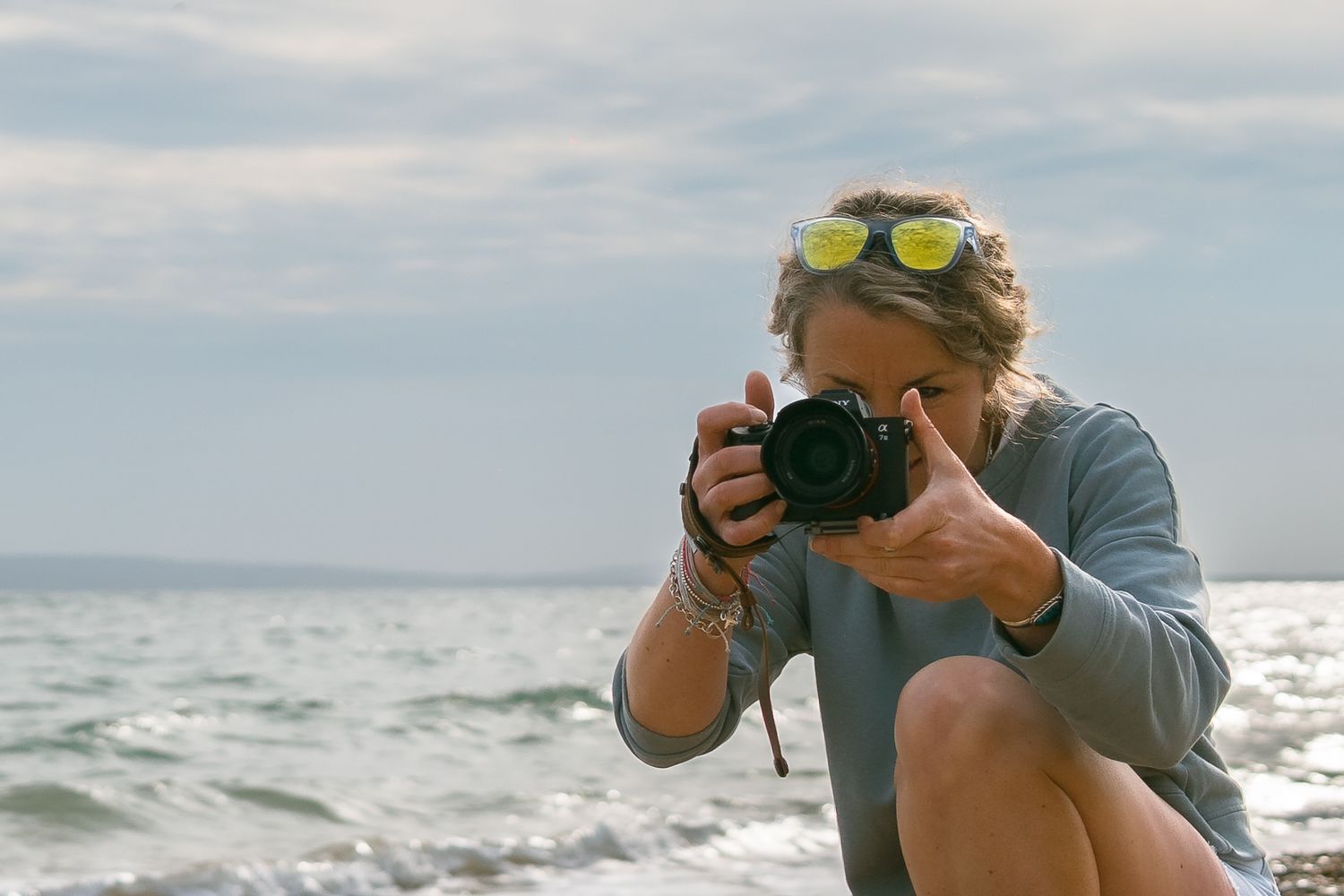 A photographer takes a series of photos on a beach while wearing a baseball cap and grey sweatshirt.