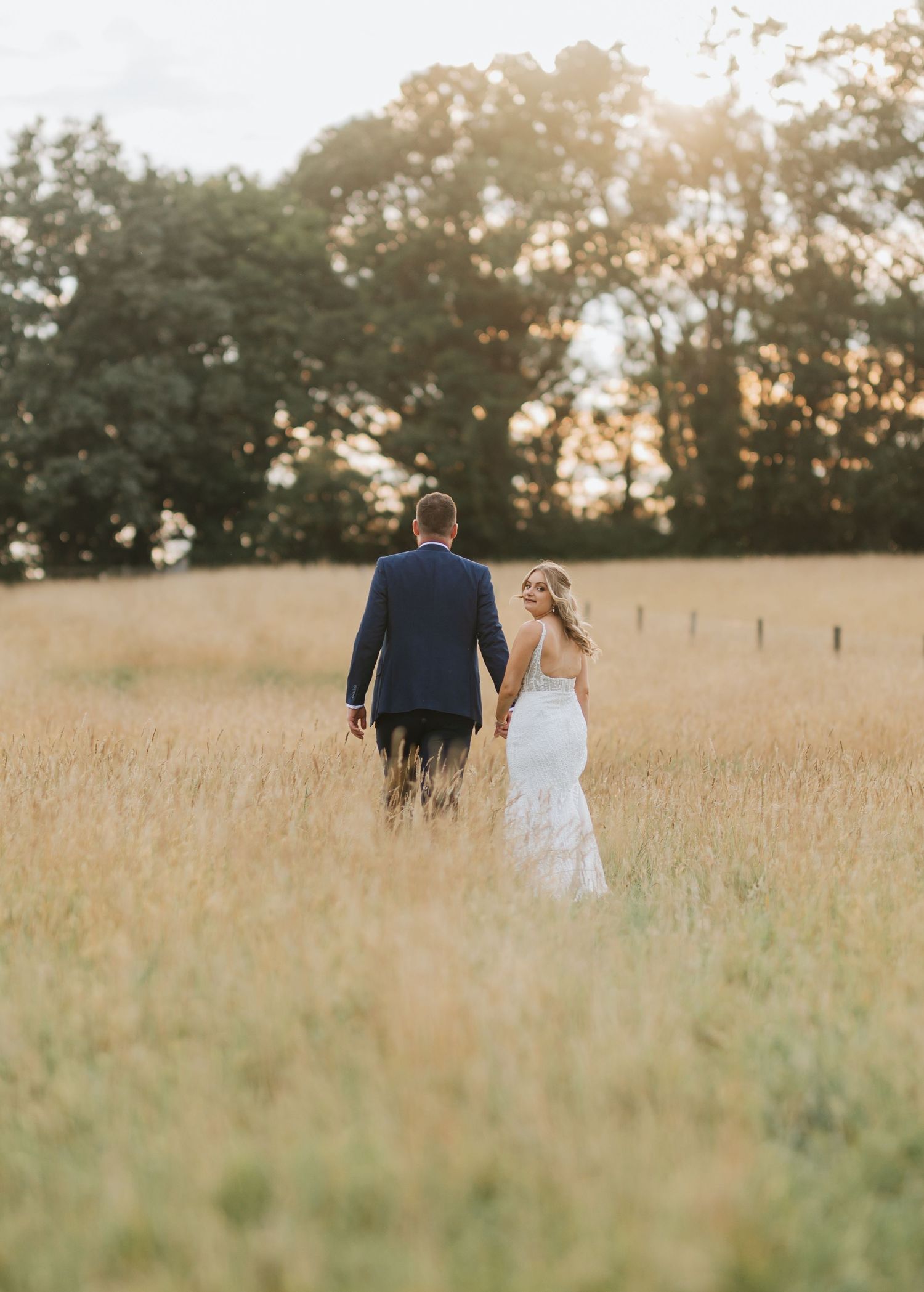A couple walks through a sunlit golden field during sunset with trees silhouetted in the background.