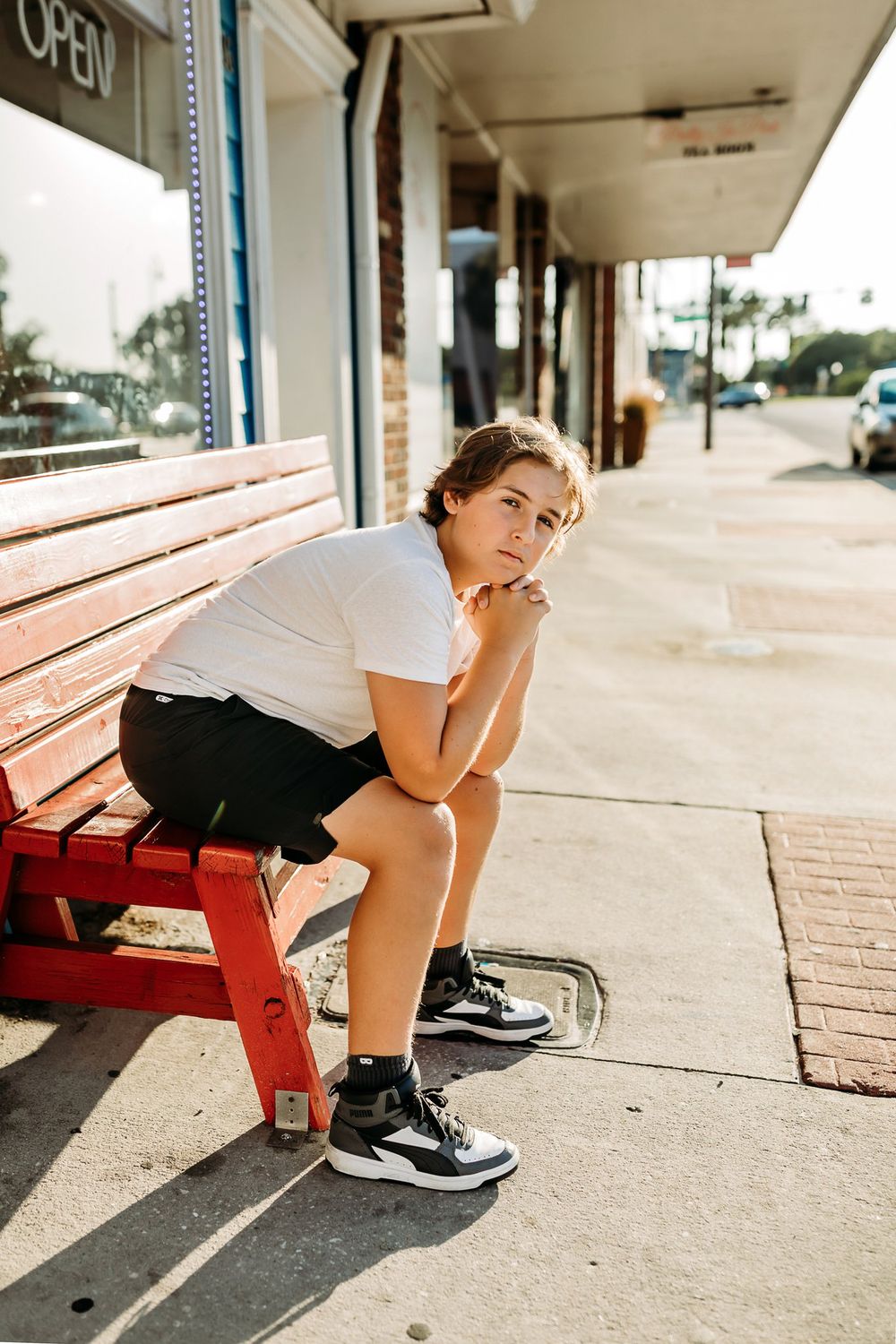 A person in athletic wear crouches down next to a red bench on a paved walkway in warm sunlight.