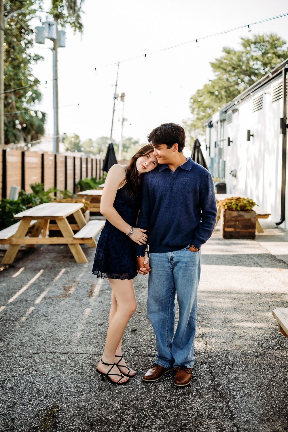 Couple embraces on gravel path with picnic tables and white buildings in background.