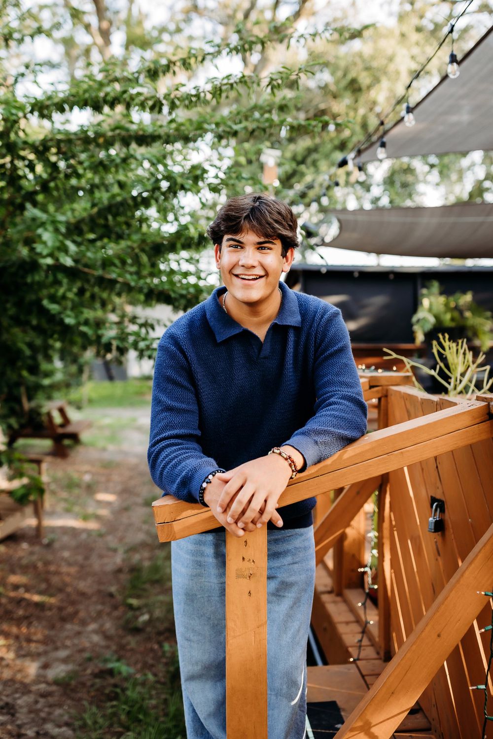 Young person in a navy blue sweater and jeans sits casually on a wooden outdoor bench on a sunny day.