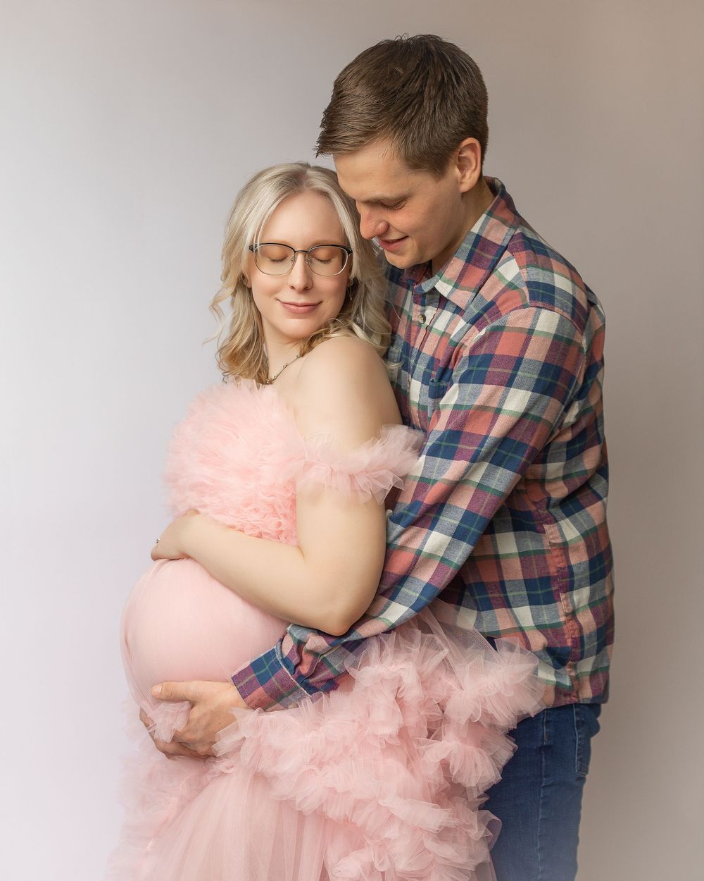 Romantic maternity shoot series features couple in plaid and pink tulle dress embracing against neutral backdrop.