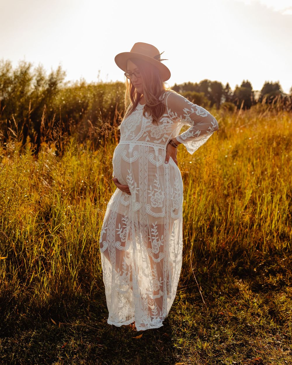 A figure wearing a white lace dress and hat stands in a golden field at sunset creating a dreamy silhouette.