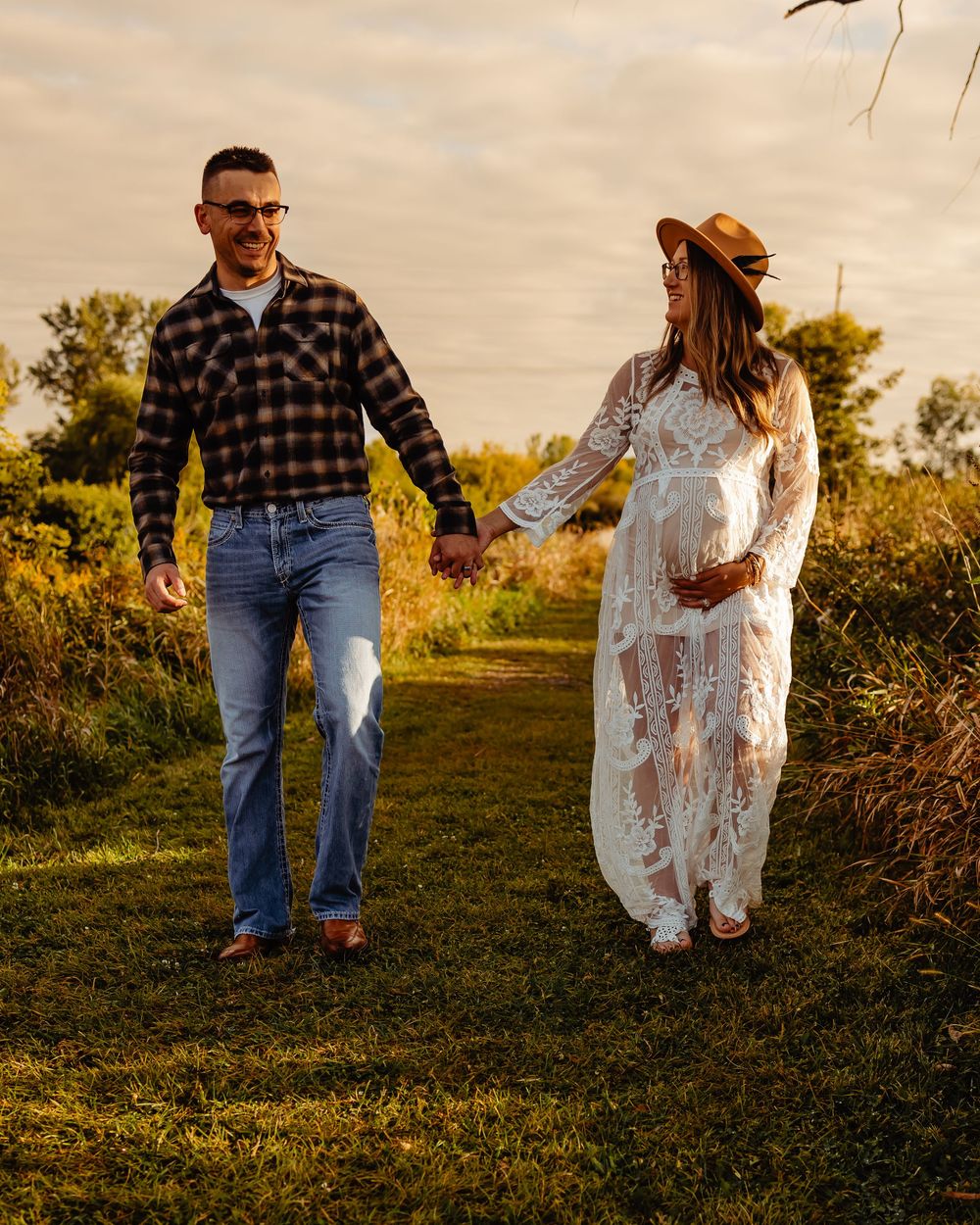 A couple walks hand in hand through a golden field at sunset while wearing casual attire and western boots.