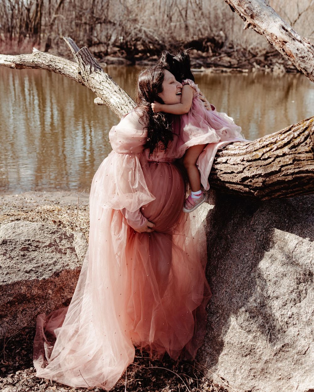 A flowing peach-colored tulle dress adorns a figure posing gracefully by a fallen tree near a tranquil pond.