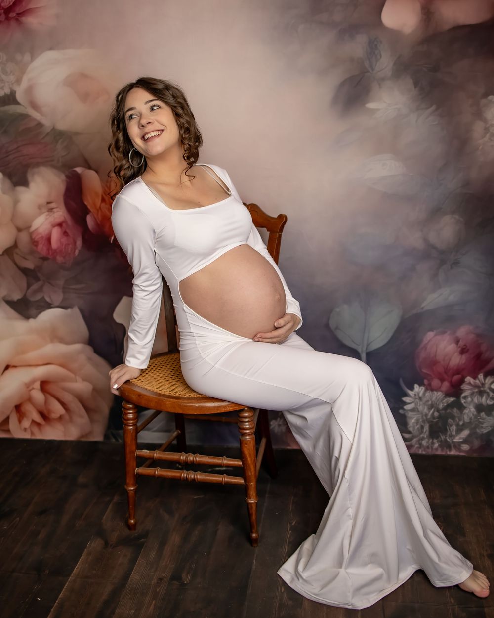 Pregnant woman in white dress poses on wooden chair in moody floral studio setting during maternity photoshoot.