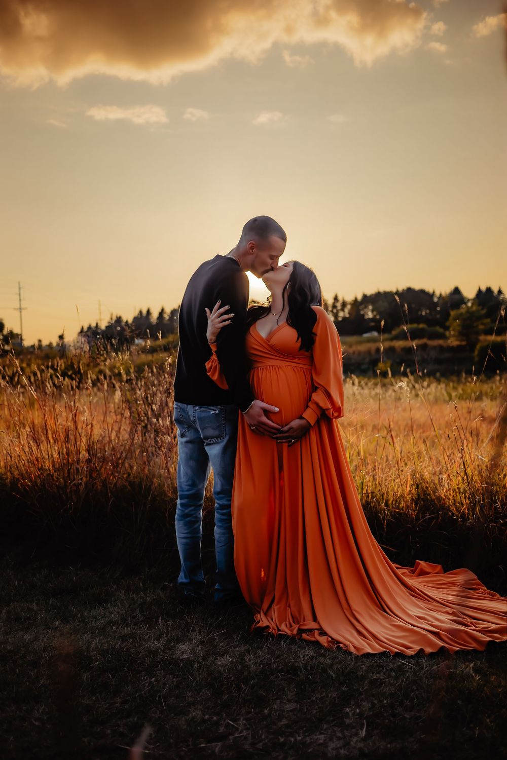 A couple embraces in a golden field during sunset, with the woman wearing a flowing burnt orange dress.