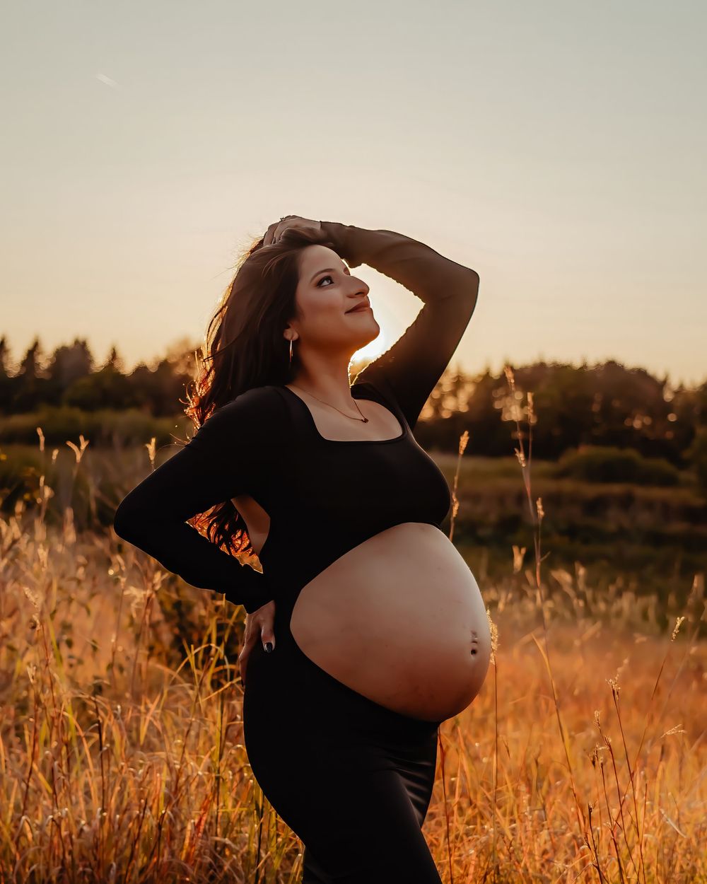 Silhouetted figure in a black dress stands in a golden field during sunset creating a warm, ethereal maternity portrait.