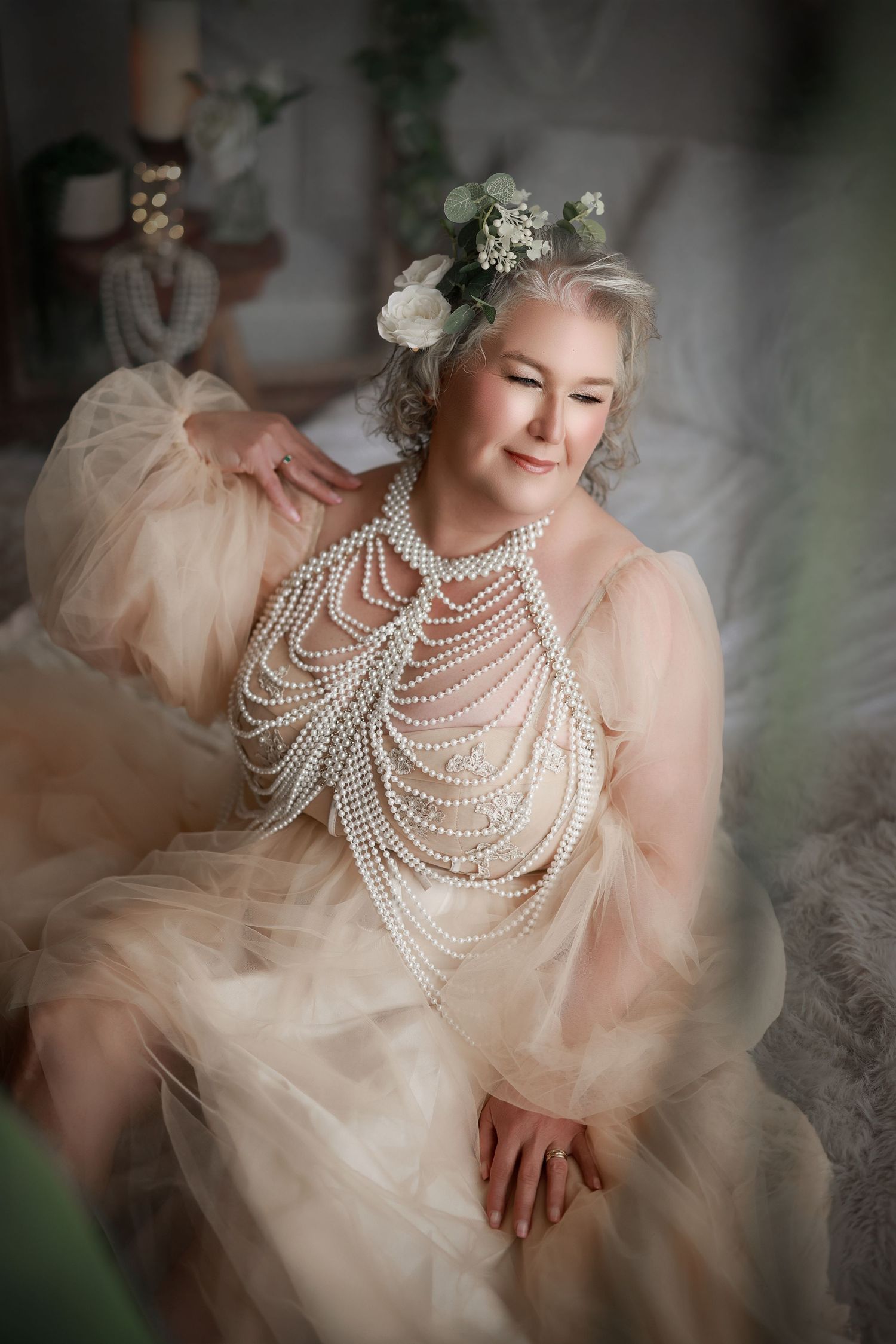 Elegant older woman in champagne tulle gown adorned with pearl necklaces and floral crown poses gracefully indoors.