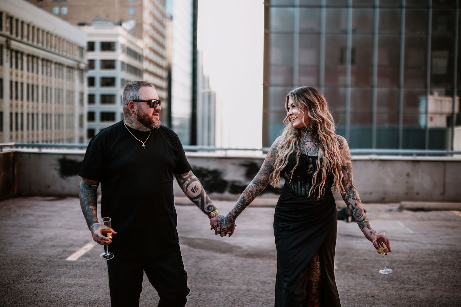 Tattooed couple holding hands walking on urban rooftop, dressed in black outfits, with city buildings in background.