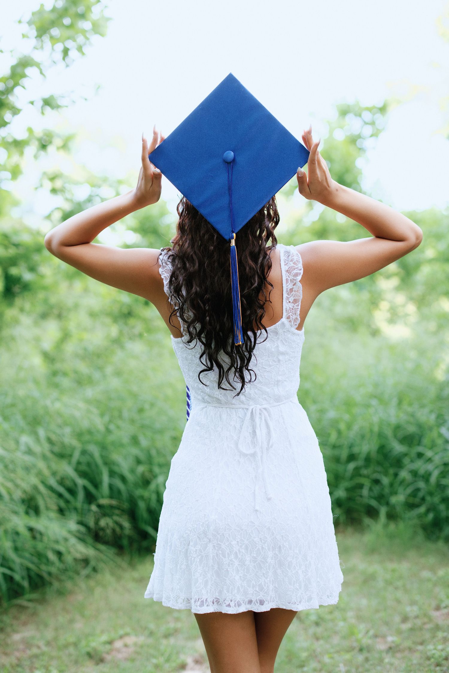 Young woman in white lace dress holding blue graduation mortarboard cap, standing outdoors among green trees.