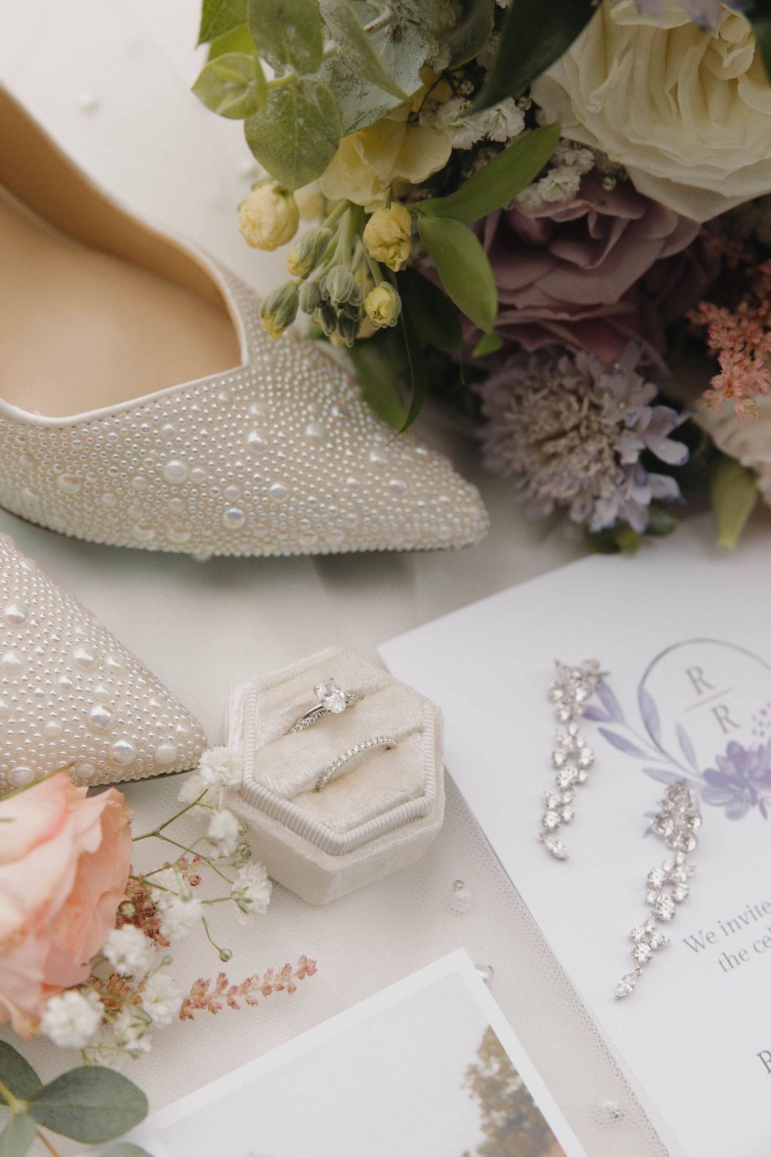 Wedding accessories including crystal shoes, floral bouquet, jewelry and invitation cards arranged on white surface.