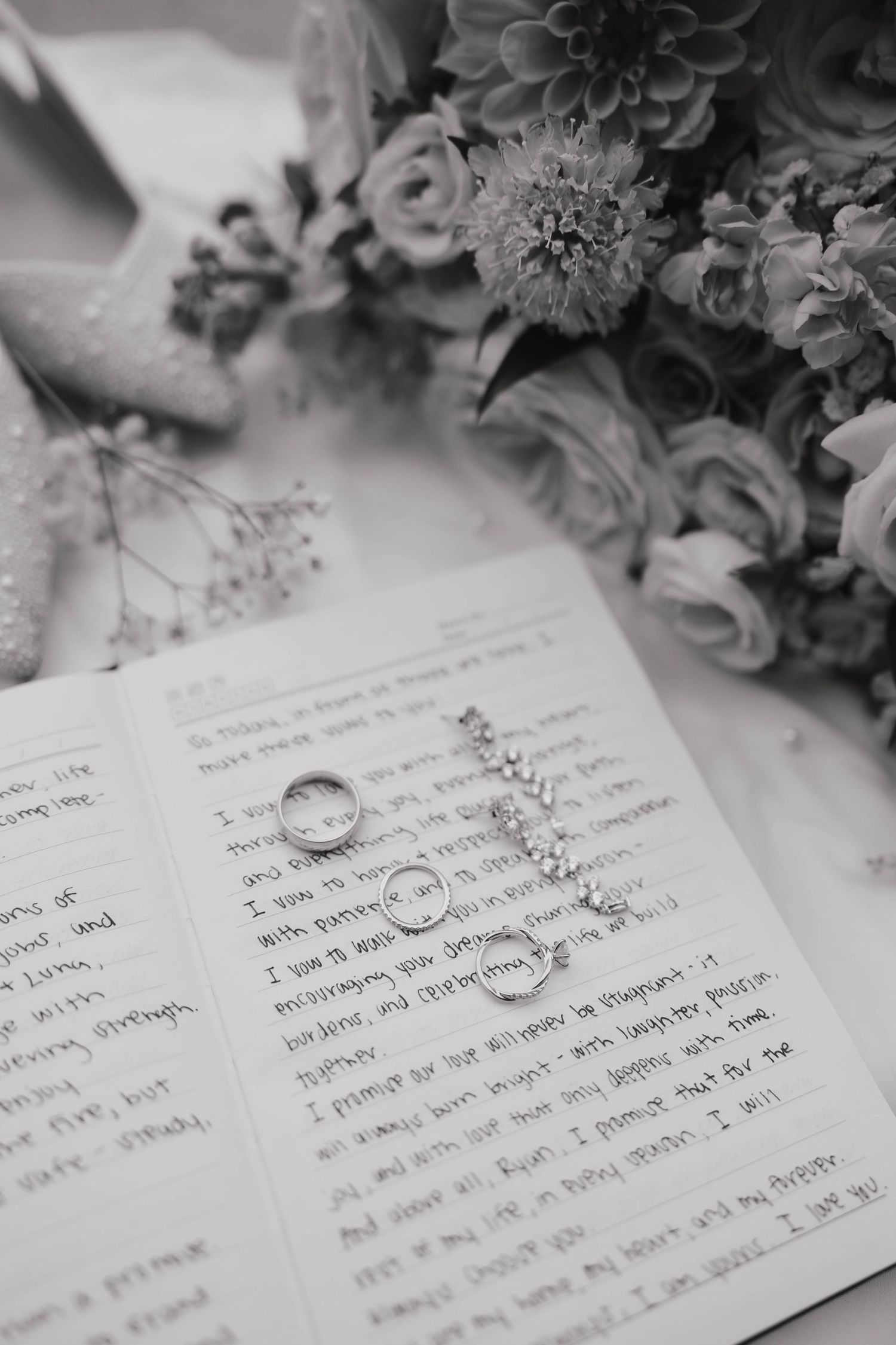 Wedding rings rest on handwritten vows with flowers in soft black and white photography.