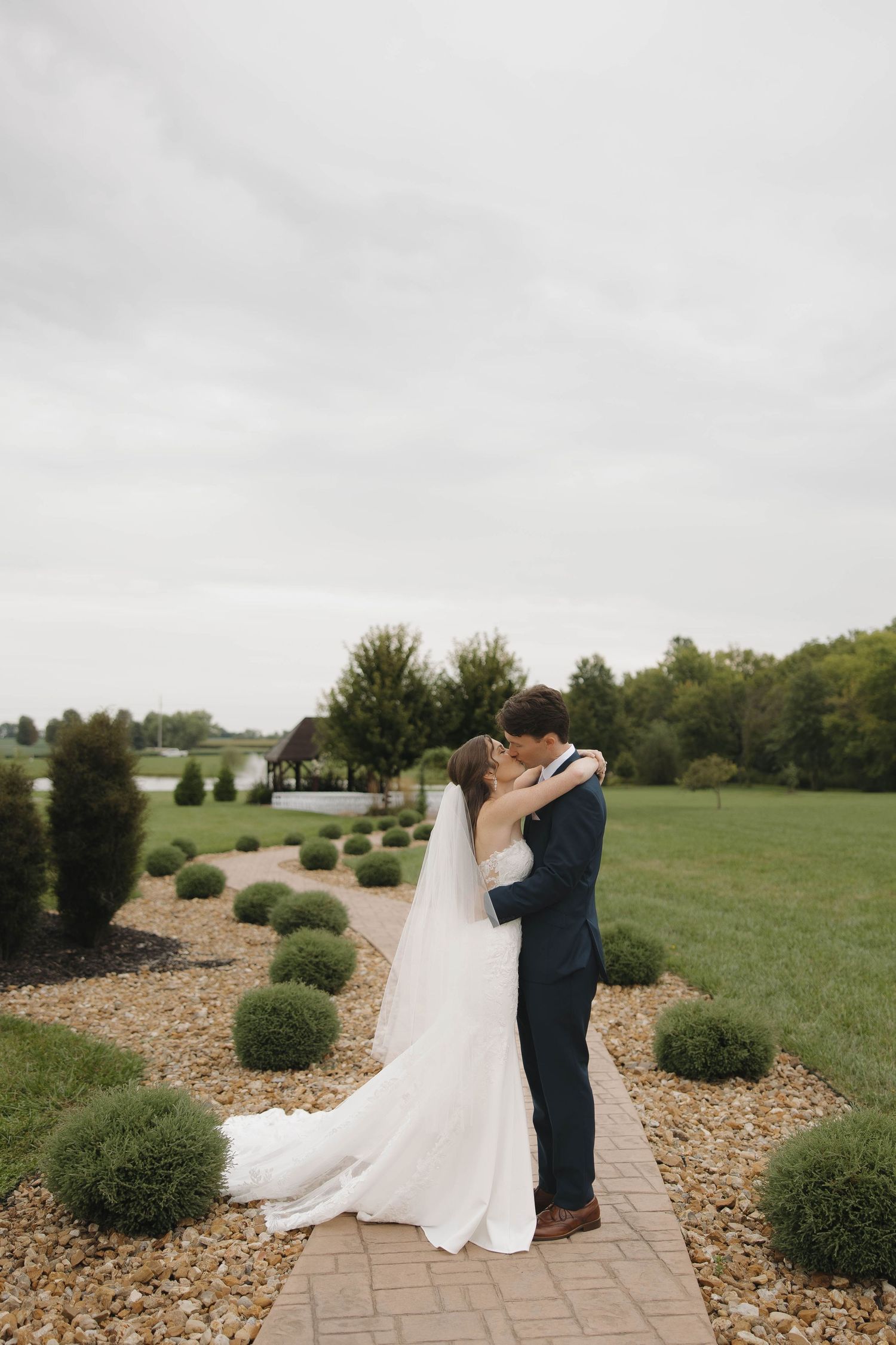 A couple in wedding attire share an intimate moment on a stone pathway surrounded by manicured shrubs.