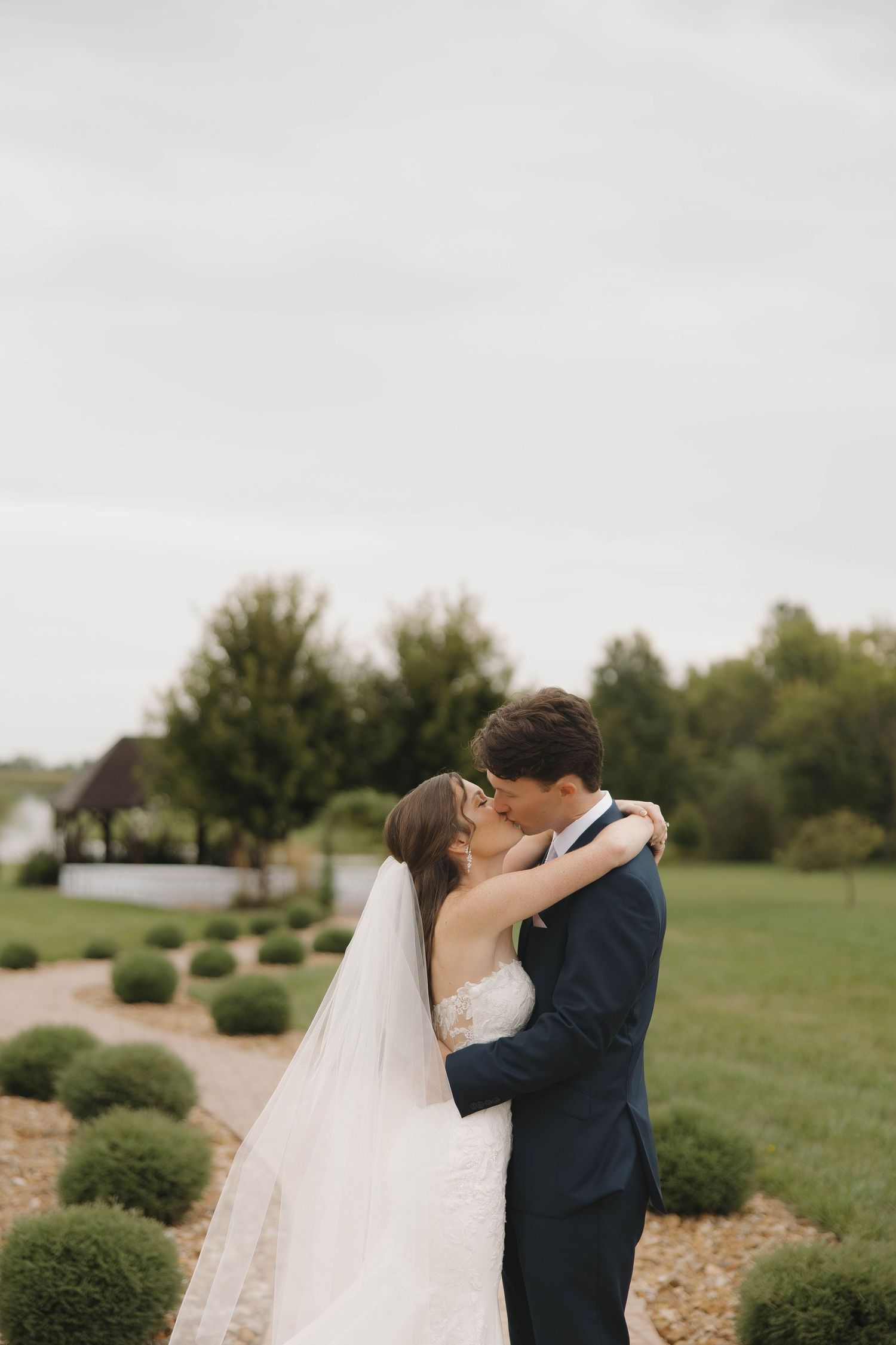 A romantic wedding couple shares embraces in a serene outdoor setting with green grass and bushes.