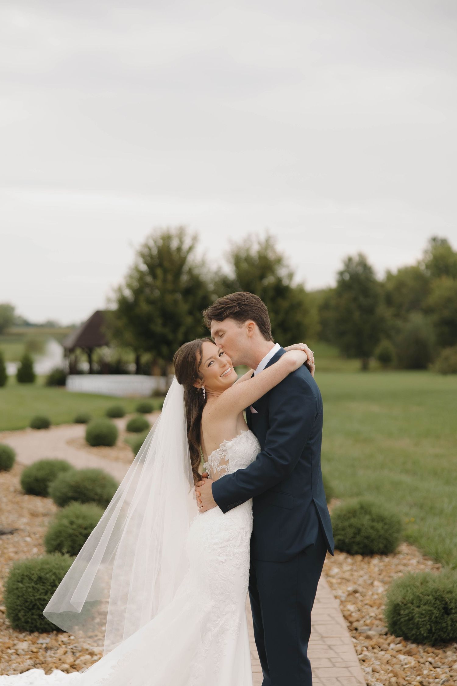 A romantic wedding couple embraces lovingly in front of green landscaping on their special day.