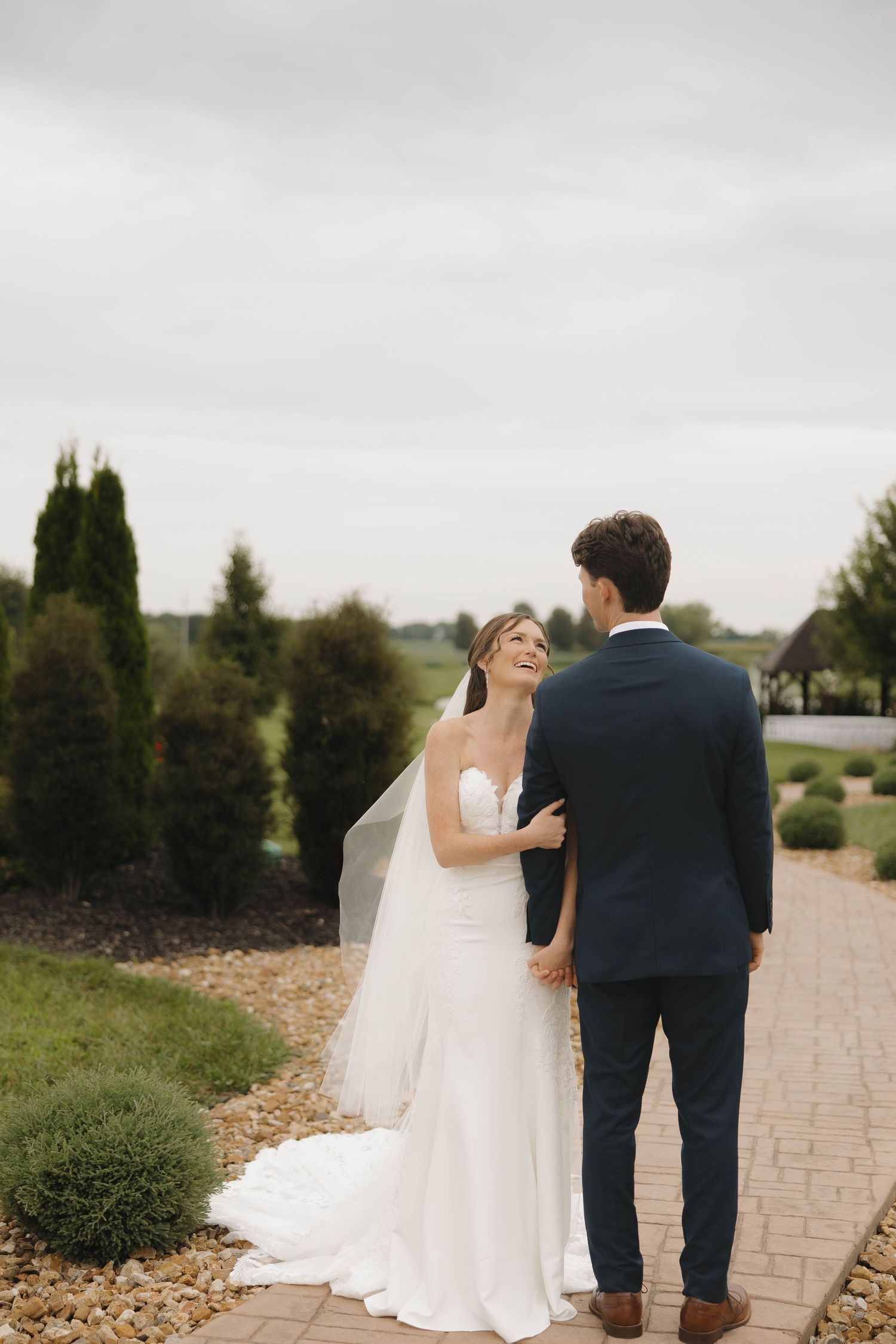 Wedding couple walks down stone path in romantic outdoor setting with lush greenery and trees in background.