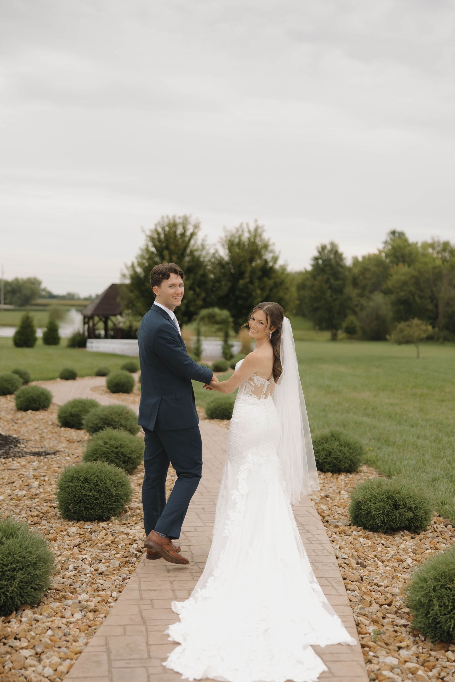Newlyweds pose on a stone path surrounded by landscaped gardens and green lawns on their wedding day.