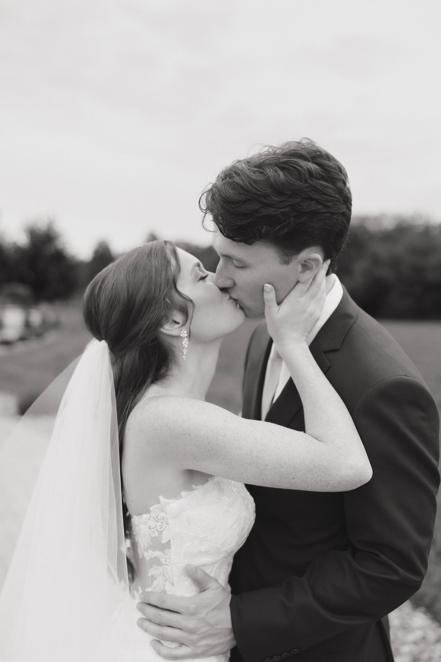 A bride and groom share a romantic kiss outdoors in black and white.