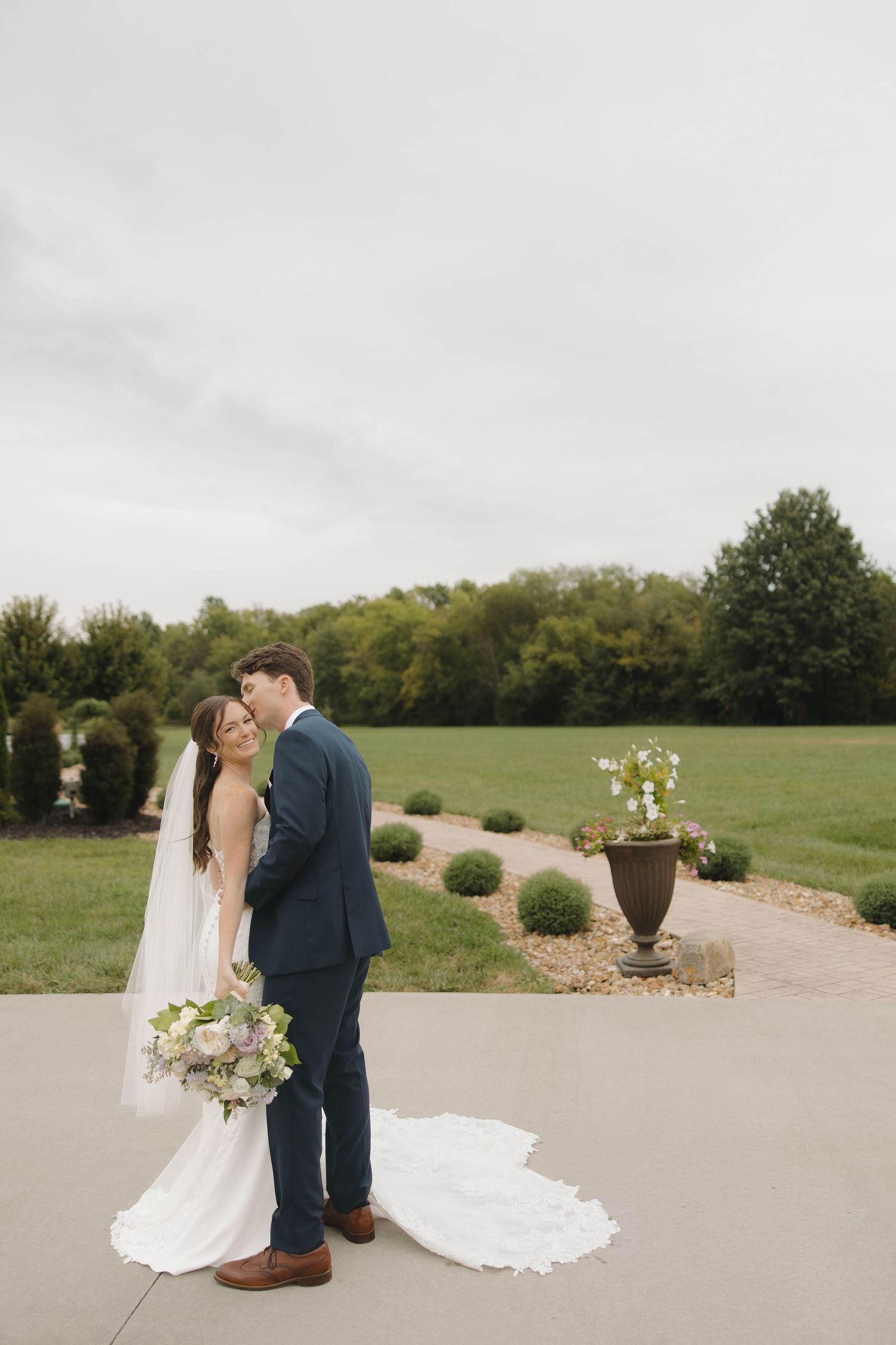 Wedding couple poses on a concrete path with lush green backdrop and decorative planters.