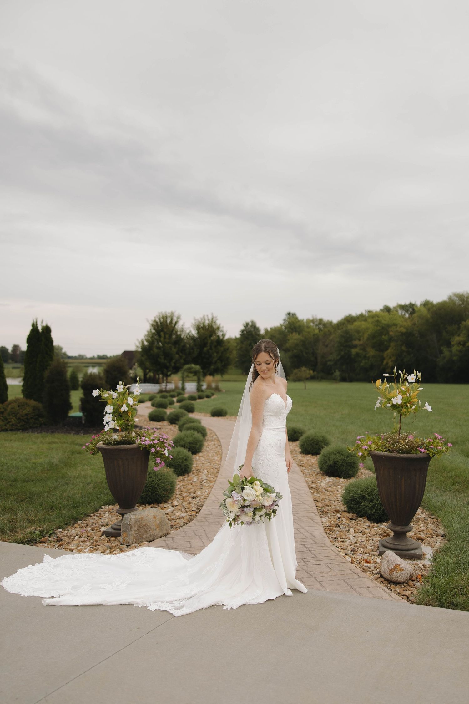 Bride in flowing white dress and veil poses along stone pathway with decorative planters and green landscape.