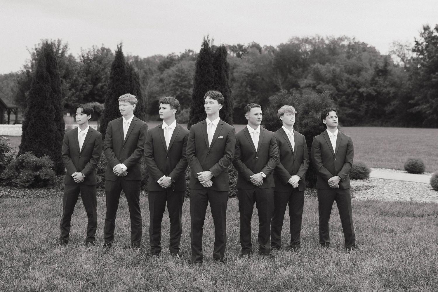 Groomsmen in dark suits line up for a formal wedding photo against a natural outdoor backdrop.