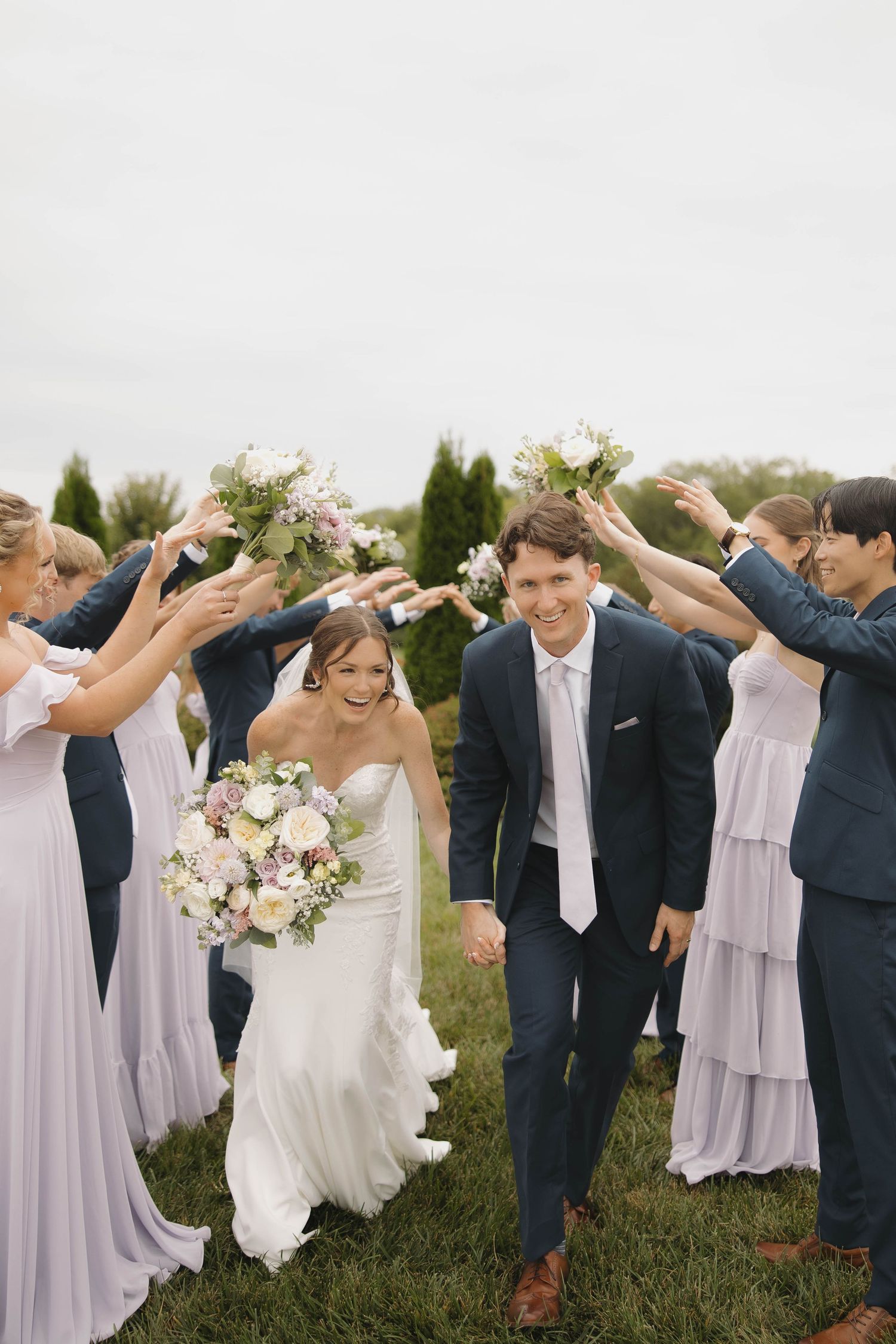 Wedding party forms a tunnel on grassy field with bridesmaids in lavender dresses and groomsmen in navy suits.